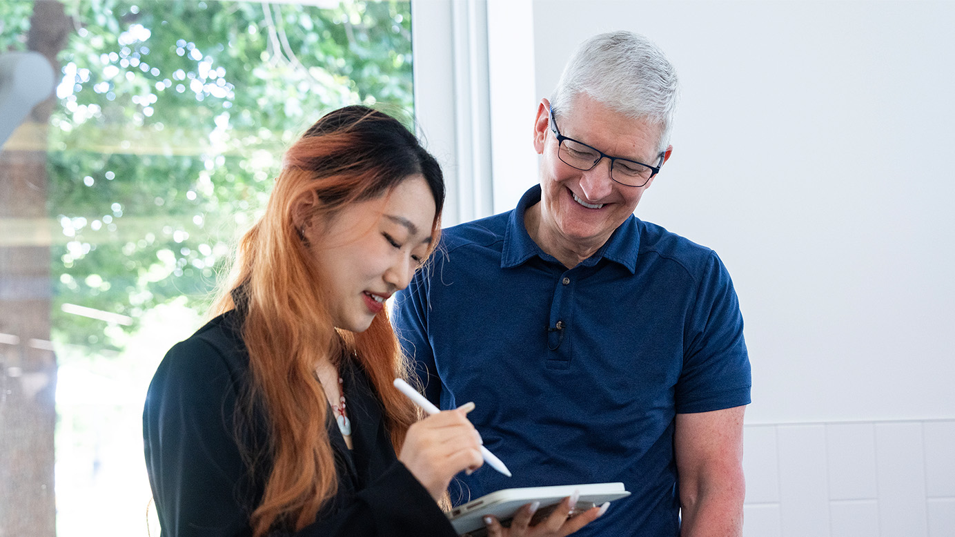 Tim Cook lächelt, während eine Gewinnerin der Swift Student Challenge bei der WWDC ein iPad verwendet.