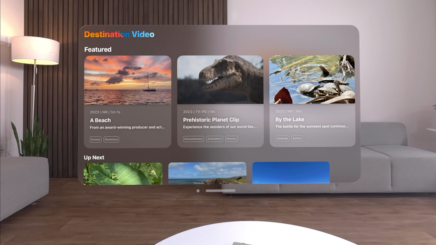 A screenshot of the Destination Video app on Apple Vision Pro. The screenshot appears front and center in a living room with white walls, gray couches, and a dark wood floor. The Destination Video app shows a section titled Featuring that contains three video options: A Beach, Prehistoric Planet Clip, and By the Lake.