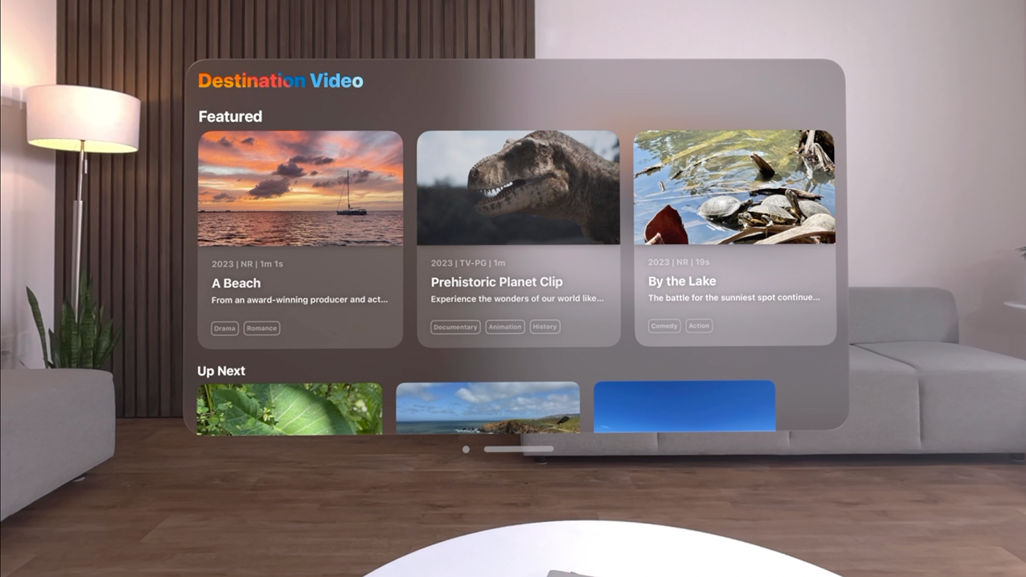 Apple Vision ProのDestination Videoアプリのスクリーンショット。白い壁、グレーのソファ、ダークウッドの床のあるリビングルームの前面中央にスクリーンショットが表示されている。「ビーチ」、「先史時代の惑星クリップ」、「湖畔」という3つのビデオオプションと、「特集」というタイトルのセクションが表示されたDestination Videoアプリ。